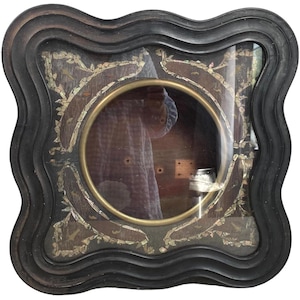 May include: A black wooden frame with a wavy design. The frame has a circular opening with a glass pane and a brass ring around the edge. The frame is decorated with small pieces of mother-of-pearl.