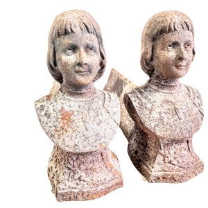 Pode incluir: Um par de suportes de lareira antigos de ferro fundido em forma de busto de uma menina. Os suportes estão desgastados e têm um aspeto rústico.