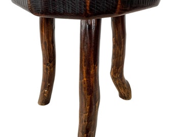Puede incluir: Un taburete de madera rústico con una tapa redonda de color marrón oscuro y tres patas robustas. La tapa tiene una superficie ligeramente irregular y está decorada con cuatro detalles metálicos.