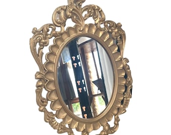 May include: A gold-toned oval mirror with a heavily ornate frame. The frame is detailed with intricate scrollwork and floral motifs.
