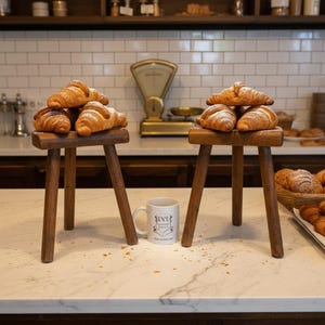 May include: Two small wooden stools each hold a stack of golden-brown croissants. A white mug with text sits on the marble countertop. A vintage scale and shelves with jars are in the background. The scene evokes a bakery or cafe.