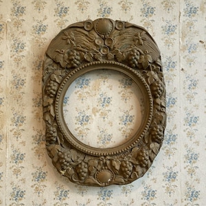 May include: Ornate, oval-shaped, antique picture frame with a bronze finish. The frame features detailed relief carvings of grapes, leaves, and decorative elements. The center is a circular opening. The frame is mounted on a floral wallpaper background.