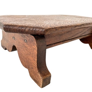 Puede incluir: Un pequeño taburete de madera con una parte superior rectangular y cuatro patas. El taburete está hecho de madera marrón oscura y tiene un diseño simple y rústico.