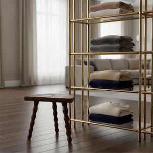May include: A three-legged wooden stool with a dark brown finish and wavy, turned legs stands beside a tall, gold-toned metal shelving unit. The shelves hold folded stacks of jumpers in shades of beige, grey, navy blue, and cream.