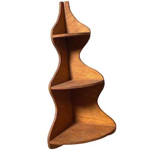 Puede incluir: Estantería esquinera de madera con un diseño único y curvo. La estantería tiene tres niveles y está hecha de madera de color marrón claro. La forma general recuerda a un reloj de arena o una figura estilizada. Diseñada para una esquina.