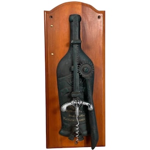 Op de afbeelding: Een vintage flessenopener van gietijzer in de vorm van een wijnfles, gemonteerd op een houten plaat. De opener heeft een zilverkleurige kurkentrekker en een tandwielmechanisme. De plaat heeft de tekst "Saur. Fr. Beaujolais Blancs et Roses".