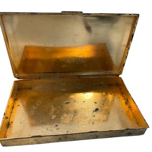 May include: An empty, gold-colored metal box with a hinged lid. The box is open and the inside is visible. The box has a worn, aged appearance.