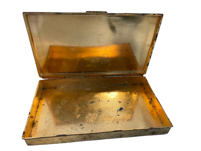 May include: An empty, gold-colored metal box with a hinged lid. The box is open and the inside is visible. The box has a worn, aged appearance.