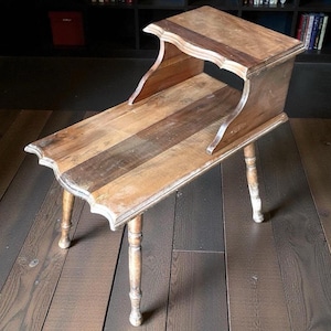 May include: A vintage wooden side table with a distressed finish. The table features a rectangular top with a scalloped edge and a lower shelf. It has four turned legs and is constructed from various shades of wood.