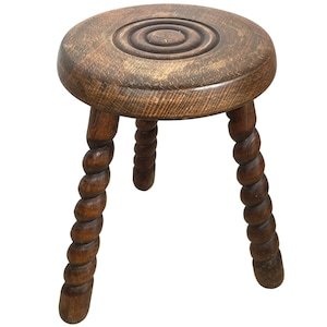 May include: A wooden stool with a round, carved top and three twisted legs. The stool is brown and has a rustic appearance.