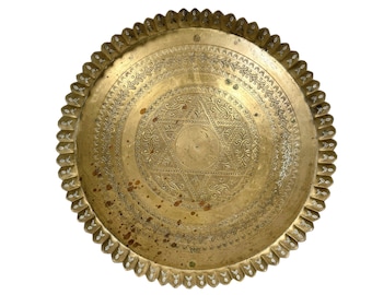 May include: A round, brass serving tray with an intricate, embossed design. The tray has a scalloped edge and a central starburst pattern.