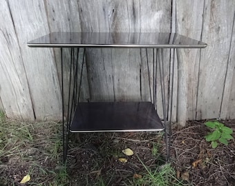 May include: A vintage black side table with a rectangular top and a lower shelf. The table has a black metal frame with thin, angled legs. The table is approximately 61 cm long, 41 cm wide, and 51 cm tall.