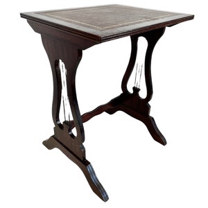 Puede incluir: Una mesa auxiliar de madera oscura con una tapa de cuero. La mesa tiene una base en forma de lira con detalles intrincados.