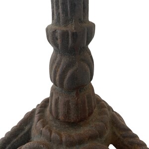 May include: Close-up of the base of a cast iron candle holder. The base is a three-pronged design with a rope-like pattern. The holder is a dark brown colour with rust spots.