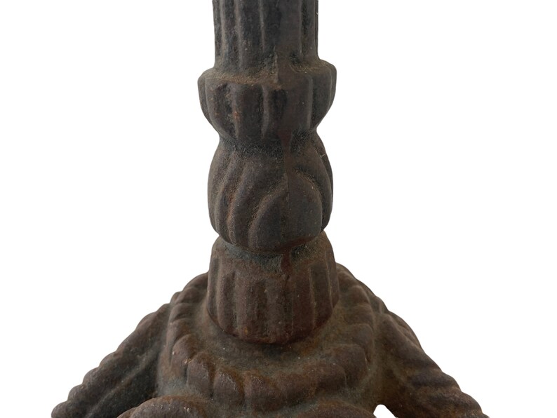 May include: Close-up of the base of a cast iron candle holder. The base is a three-pronged design with a rope-like pattern. The holder is a dark brown colour with rust spots.