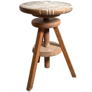 May include: A wooden adjustable stool with a round seat and three legs. The stool has a white paint splatter on the seat.