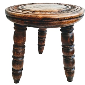 May include: A small, round, wooden stool with three turned legs. The top of the stool has a carved design.