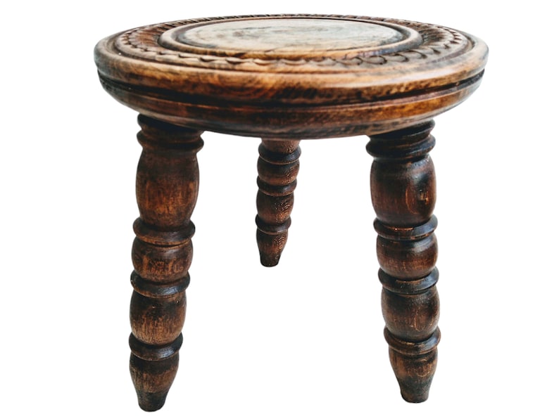May include: A small, round, wooden stool with three turned legs. The top of the stool has a carved design.