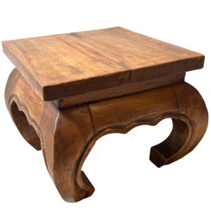 May include: A small, square wooden table with a warm brown finish. The tabletop is flat, supported by a base with curved, decorative legs. The table is likely used for display or as a small accent piece.