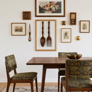 May include: A dining set featuring a rectangular wooden table and two chairs with patterned olive and brown upholstery. The wall displays a collection of framed art, a wooden clock, a brass sphere, and a large wooden spoon and fork wall hanging.