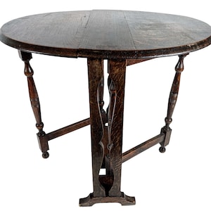 Op de afbeelding: Een donkerbruine houten klaptafel met een ronde bovenkant en een unieke X-vormige basis. De tafel heeft een verweerde afwerking en is gemaakt van massief hout.