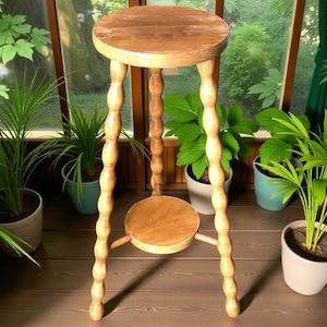 Peut inclure: Un support de plante en bois brun clair avec un dessus rond et une petite étagère ronde. Les pieds sont finement sculptés avec un motif de bobine. Le support mesure environ 76 cm de haut.