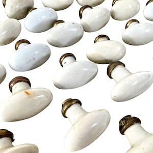 Può includere: Una collezione di pomelli per porte in porcellana bianca vintage con accenti in ottone. I pomelli sono di forma ovale e hanno una superficie leggermente testurizzata.