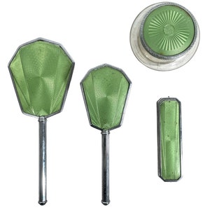 May include: A vintage green and silver vanity set with two hand mirrors and a compact. The mirrors have a geometric design and the compact has a round lid.