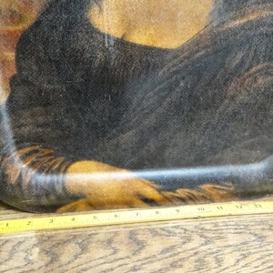 Puede incluir: Una bandeja de servir vintage con una imagen impresa de la Mona Lisa. La bandeja est&aacute; hecha de un material marr&oacute;n oscuro y tiene un borde ligeramente elevado. La imagen est&aacute; ligeramente descolorida y tiene un aspecto texturizado.