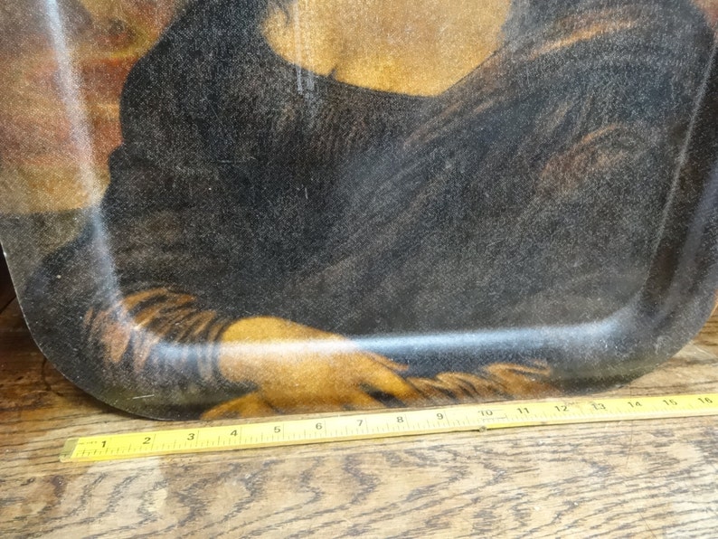 Puede incluir: Una bandeja de servir vintage con una imagen impresa de la Mona Lisa. La bandeja est&aacute; hecha de un material marr&oacute;n oscuro y tiene un borde ligeramente elevado. La imagen est&aacute; ligeramente descolorida y tiene un aspecto texturizado.