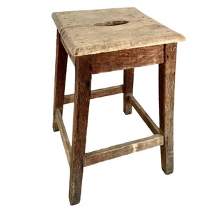 May include: A rustic wooden stool with a square top and four legs. The stool is made of dark brown wood and has a worn, distressed finish.