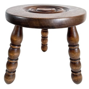May include: A dark brown wooden stool with three turned legs and a round top. The top has a concentric circle design.