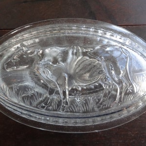May include: A clear glass butter dish with a raised design of a cow in a field of grass.