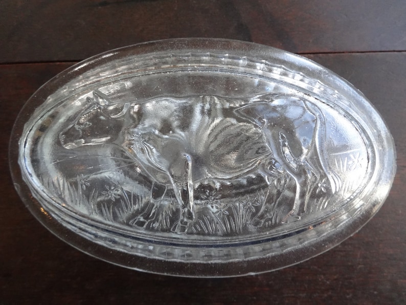 May include: A clear glass butter dish with a raised design of a cow in a field of grass.