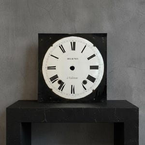 May include: Square clock face with a white dial and black Roman numerals. The clock face has the name "DELAUNEY" and the words "à Coutances" printed on it. The clock face is set against a black background.