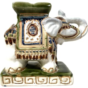 May include: A white ceramic elephant figurine with green, brown and blue painted details. The elephant is standing on a green and white base with a decorative border.
