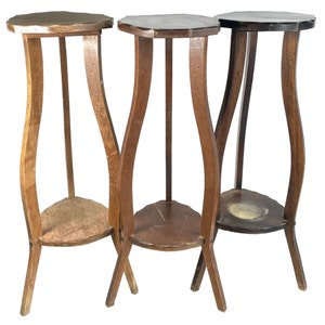 May include: Three antique wooden plant stands. Each stand has a unique, wavy leg design and a small, triangular shelf. The tops are octagonal. The wood tones vary from light to dark brown.
