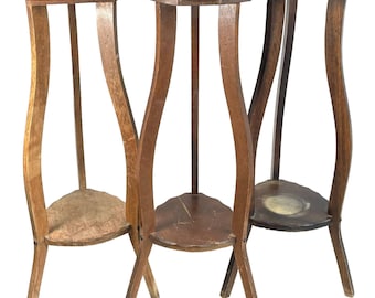 May include: Three antique wooden plant stands. Each stand has a unique, wavy leg design and a small, triangular shelf. The tops are octagonal. The wood tones vary from light to dark brown.
