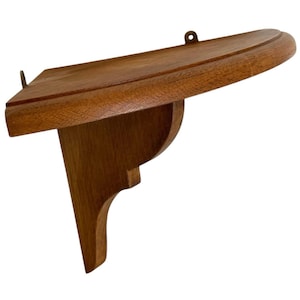 May include: A small, wooden wall shelf with a curved front edge and a decorative bracket. The shelf is made of a medium-toned wood and has a small metal loop for hanging. The shelf is approximately 10 inches wide.