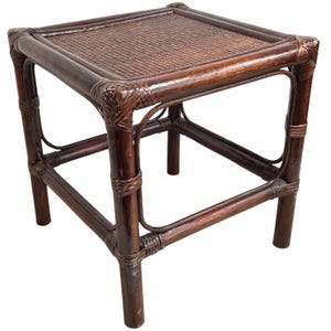 Puede incluir: Una mesa auxiliar cuadrada de ratán tejido de color marrón oscuro con una tapa tejida. La mesa tiene un diseño sencillo con cuatro patas y un estante inferior.
