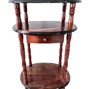 May include: A dark brown wooden side table with a round top and three shelves. The table has a drawer on the middle shelf and turned legs.