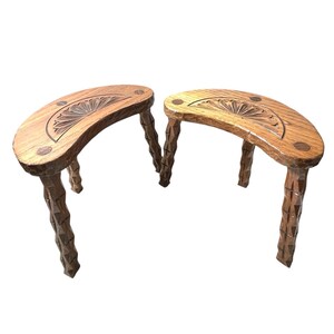 Puede incluir: Dos taburetes de madera en forma de media luna. Cada taburete tiene un diseño floral tallado en la parte superior y tres patas talladas. Los taburetes son de madera de color marrón claro y están sobre un fondo blanco.