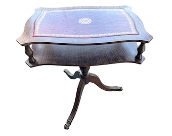 Puede incluir: Una mesa auxiliar de madera oscura con una tapa de cuero y un diseño floral decorativo. La mesa tiene un solo estante y tres patas.