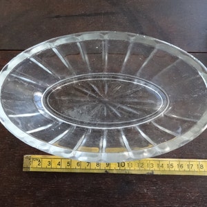 May include: A clear glass oval dish with a raised rim and a smaller oval dish inside. The dish has a decorative pattern of lines radiating from the center.