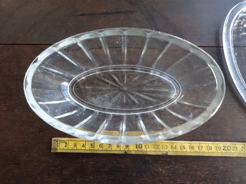 May include: A clear glass oval dish with a raised rim and a smaller oval dish inside. The dish has a decorative pattern of lines radiating from the center.