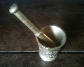 May include: A brass mortar and pestle set with an ornate design. The mortar is a bowl-shaped vessel with a wide opening and a rounded bottom. The pestle is a long, cylindrical tool with a rounded end.