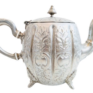 Peut inclure: Une théière en argent avec un couvercle et une anse incurvée. La théière présente un motif floral et repose sur trois petits pieds. Le bec verseur est incurvé et le couvercle est orné d'un petit bouton décoratif.