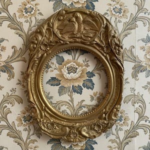 May include: Ornate gold-colored oval picture frame with detailed carvings, including birds at the top. The frame surrounds a floral patterned fabric. The frame is mounted on a floral wallpaper background.