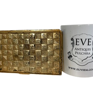 May include: A gold metal box with a geometric pattern on the top. The box is closed and has a hinged lid. The box is sitting on a white surface next to a white mug with the text "EVE ANTIQUIS PULCHRA" and "www.euviem.com".