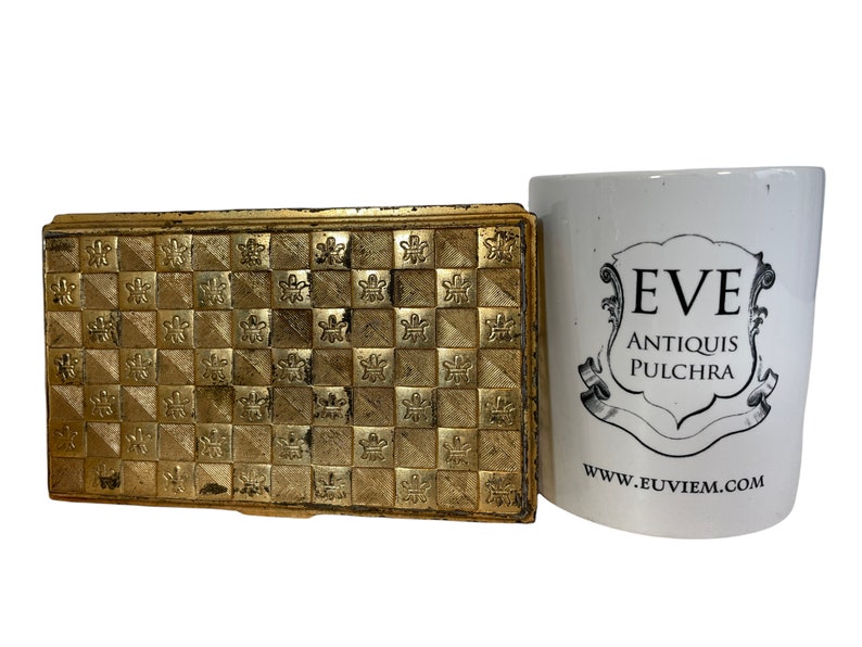 May include: A gold metal box with a geometric pattern on the top. The box is closed and has a hinged lid. The box is sitting on a white surface next to a white mug with the text "EVE ANTIQUIS PULCHRA" and "www.euviem.com".